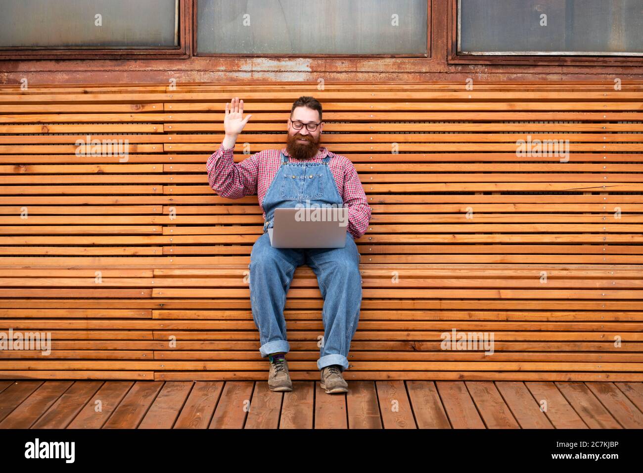Portrait of cheerful brutal bearded hipster man in blue overalls, checked shirt using on laptop talks with his girlfriend and waving his hand Stock Photo