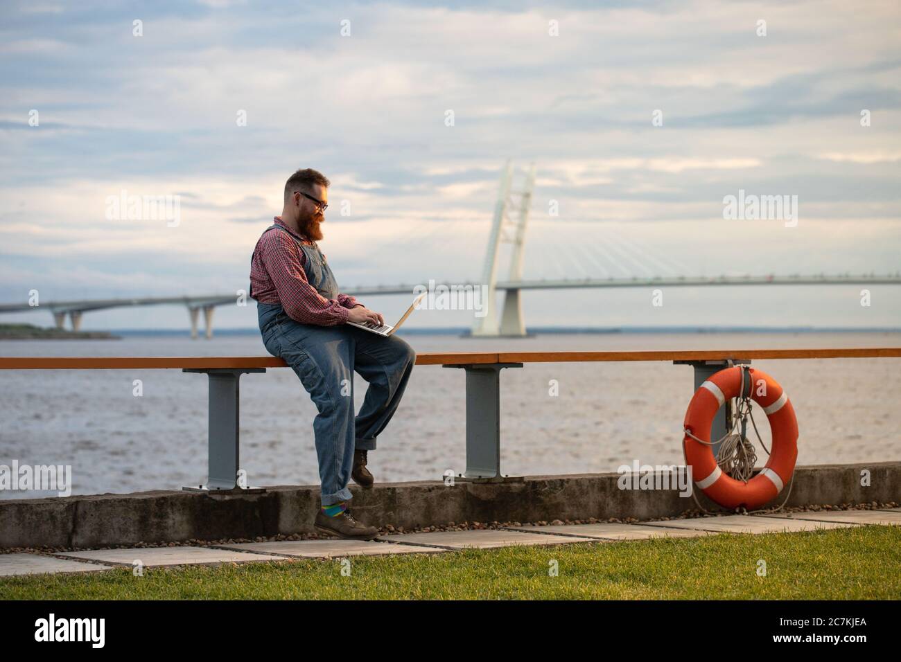 Portrait of brutal bearded man freelancer wearing blue overalls and checked shirt working on laptop at embankment outdoors. Distance job, remote work. Stock Photo