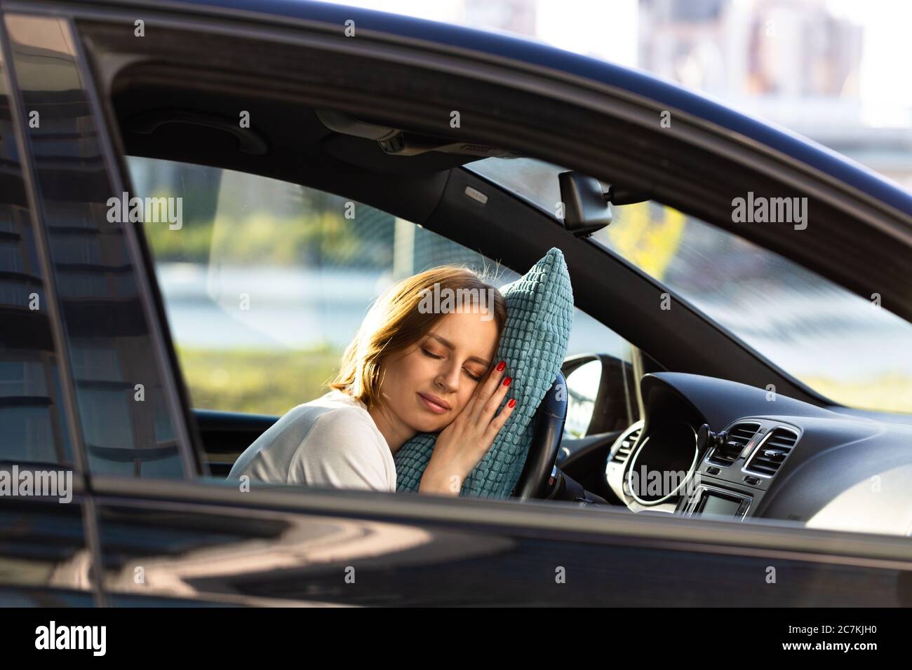 Tired young woman asleep on pillow on steering wheel, resting after long hours driving a car. Fatigue. Sleep deprivation. Stock Photo