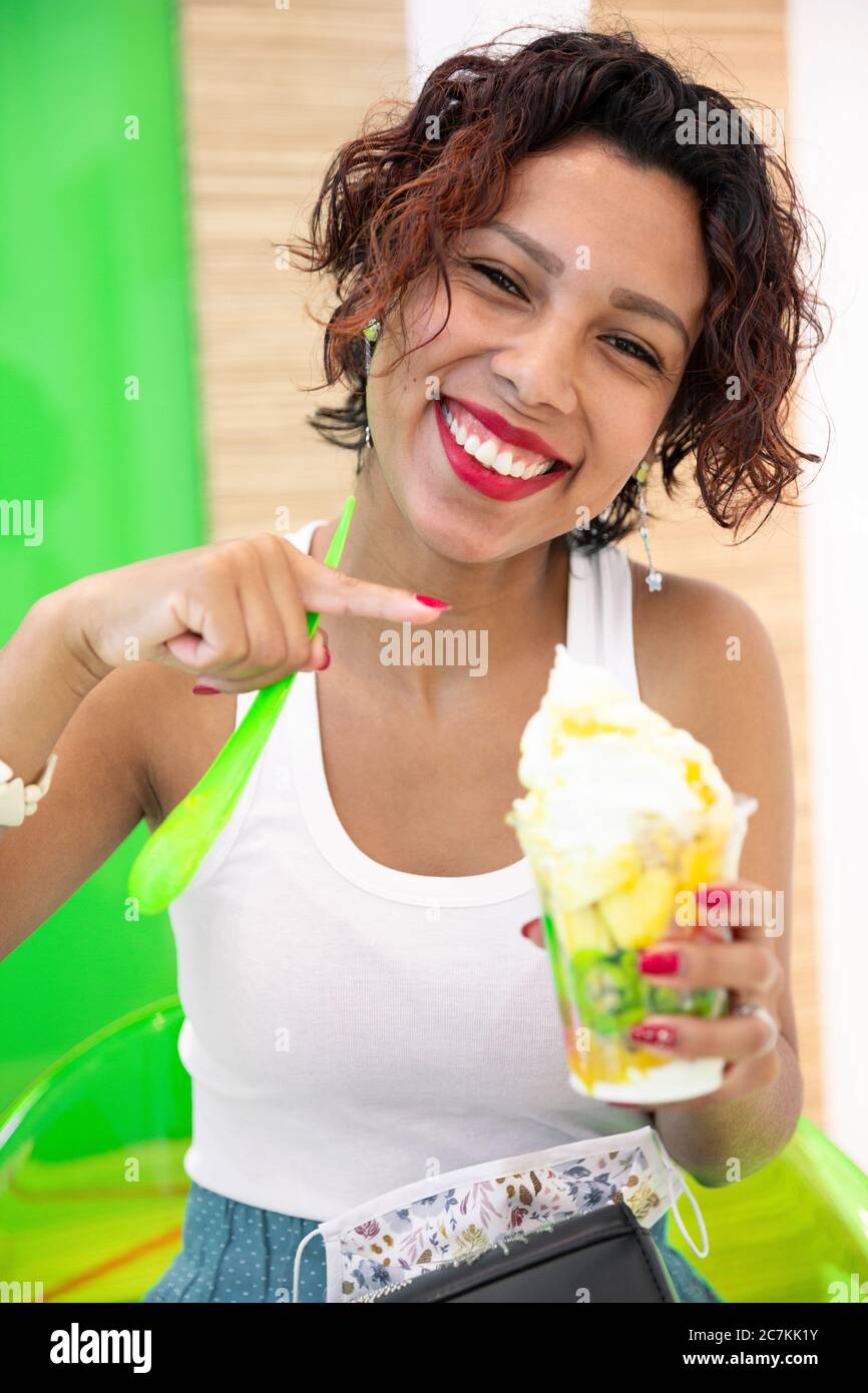 Portrait of a young woman with dark skin pointing at a glass of ice cream with her hands. She is smiling. Selective focus. Summer and lifestyle concep Stock Photo