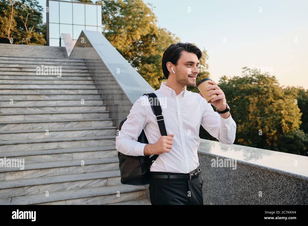 Young smiling bearded brunette man in white shirt with wireless earphones standing with cup of coffee to go in hand and backpack on shoulder happily Stock Photo