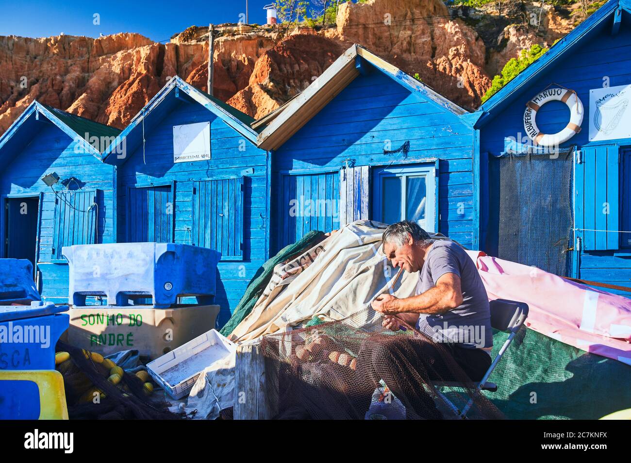 Europe, Portugal, Algarve, Litoral, Barlavento, Faro district, near Albufeira, Praia dos Olhos de Agua, fishermen mending nets, blue fishermen's huts off the red-brown cliffs Stock Photo