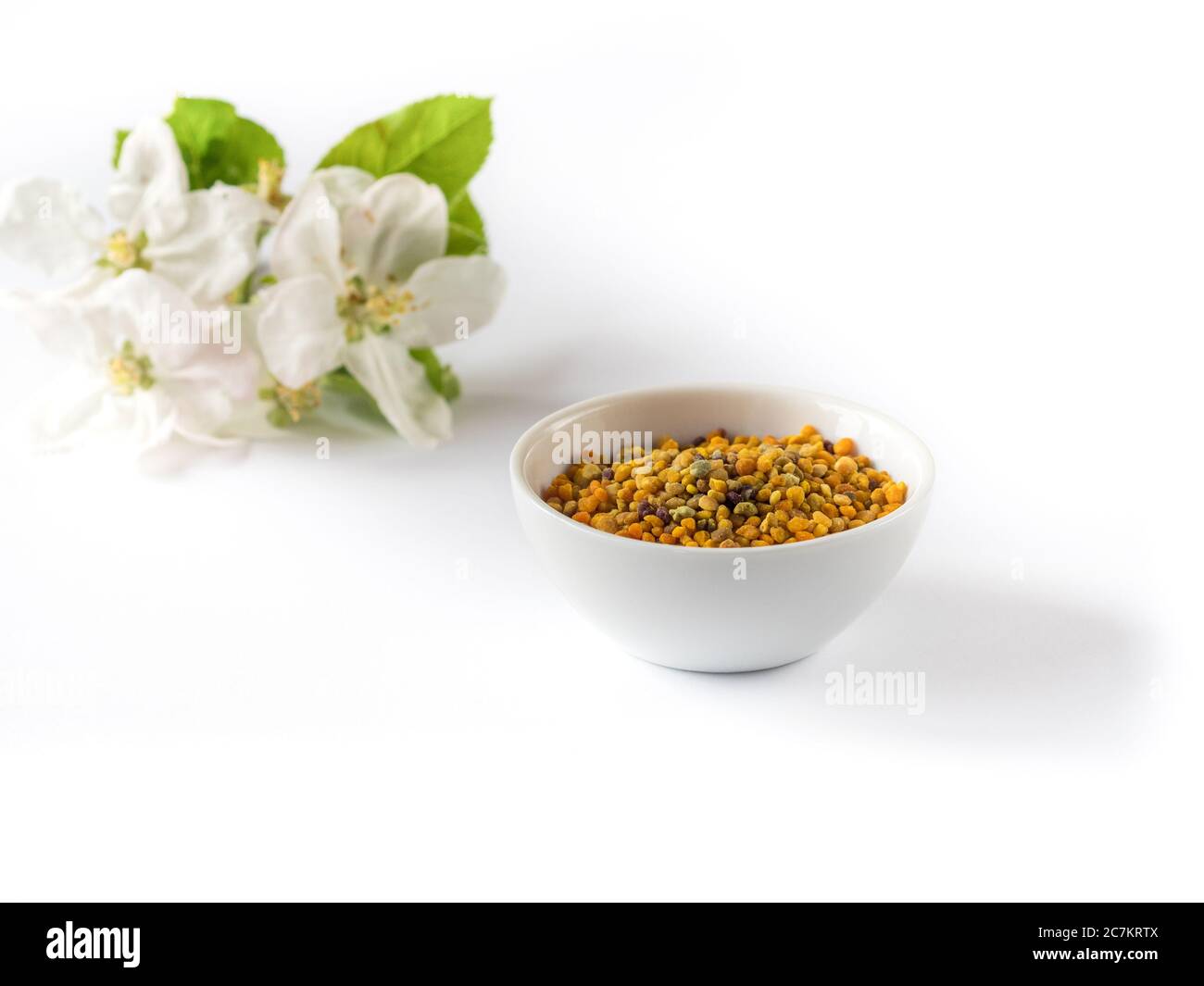 Bee pollen grains in white ceramic bowl and apple flowers on white background. Stock Photo