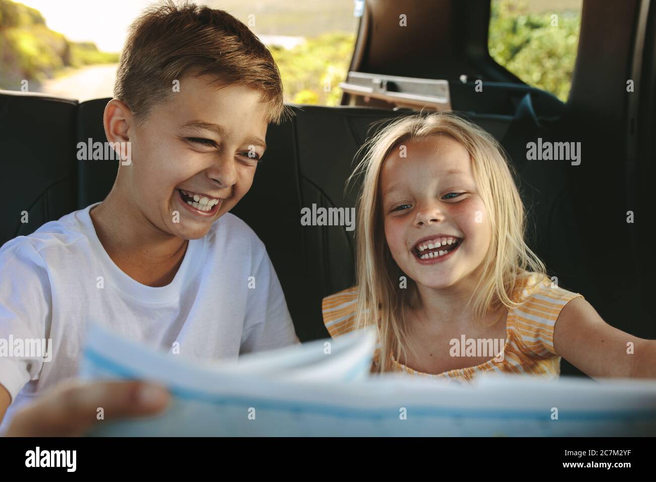 Two children sitting on backseat of car looking at map and smiling on road trip. Kids traveling in a car. Stock Photo