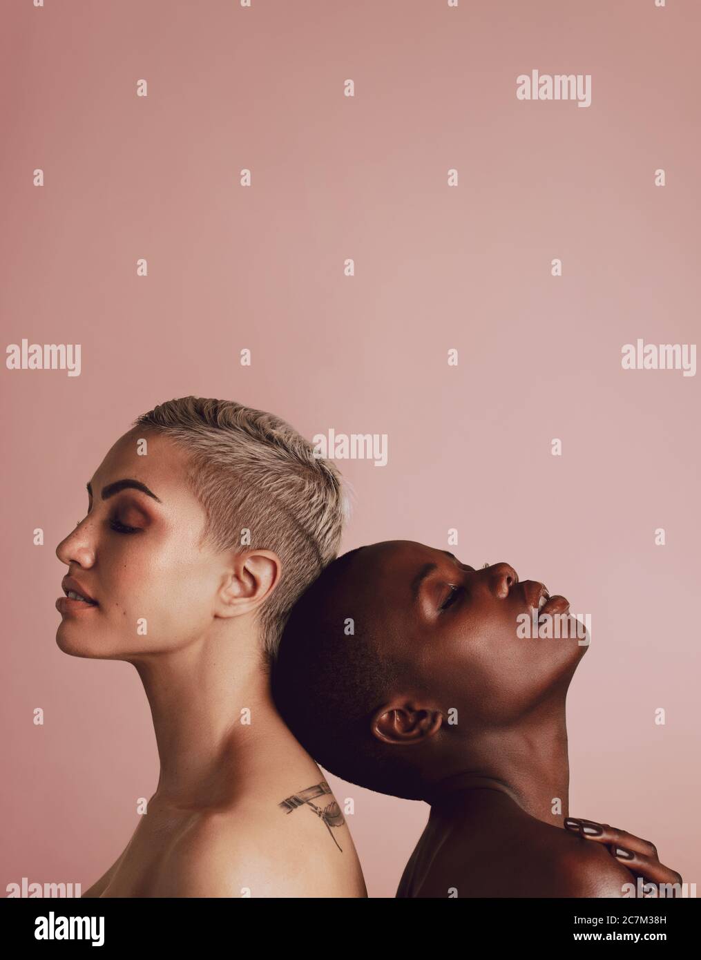 Side view of two diverse women against each other over beige background with copy space. Female models of different ethnicity standing back to back an Stock Photo