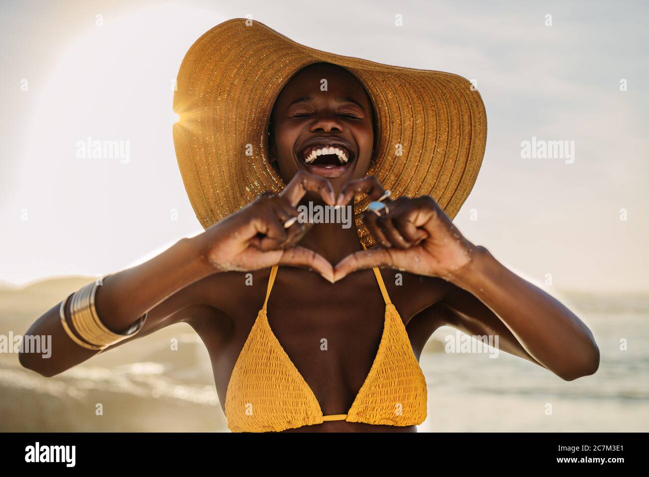 Smiling woman in bikini standing on the beach making a heart shape with her fingers. Female at the seashore wearing a sun hat making heart with hands. Stock Photo