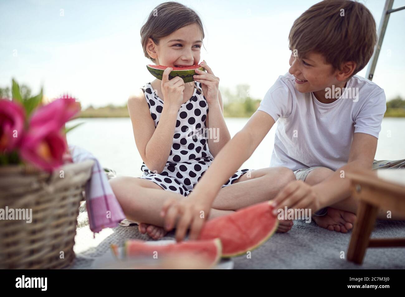 Cute kids  eating fresh watermelon on the pier.  Girl and boy enjoying together at lake. Stock Photo