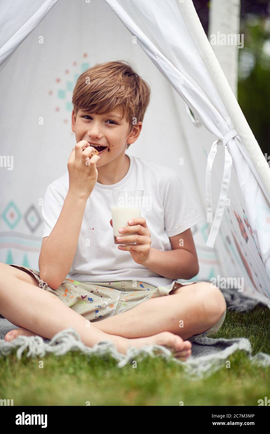 Cute boy sitting in teepee at backyard drink milk and eat cookies. Stock Photo