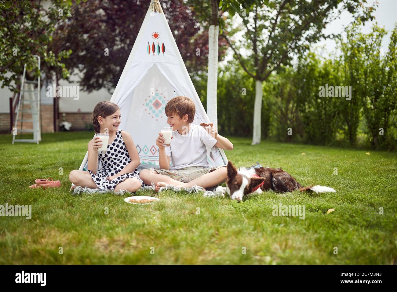 Cute happy little children sitting in teepee at backyard and drink milk. Stock Photo