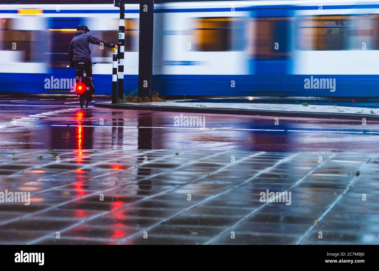 AMSTERDAM, THE NETHERLANDS JULY 9, 2020: Cyclist in the rain with a helmet on waiting at the traffic sign while a tram is passing by. Low angle and mo Stock Photo