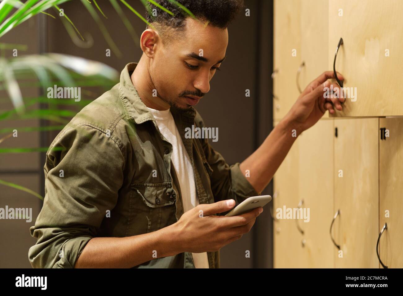 Side view of young attractive African American guy dreamily using cellphone in locker room Stock Photo