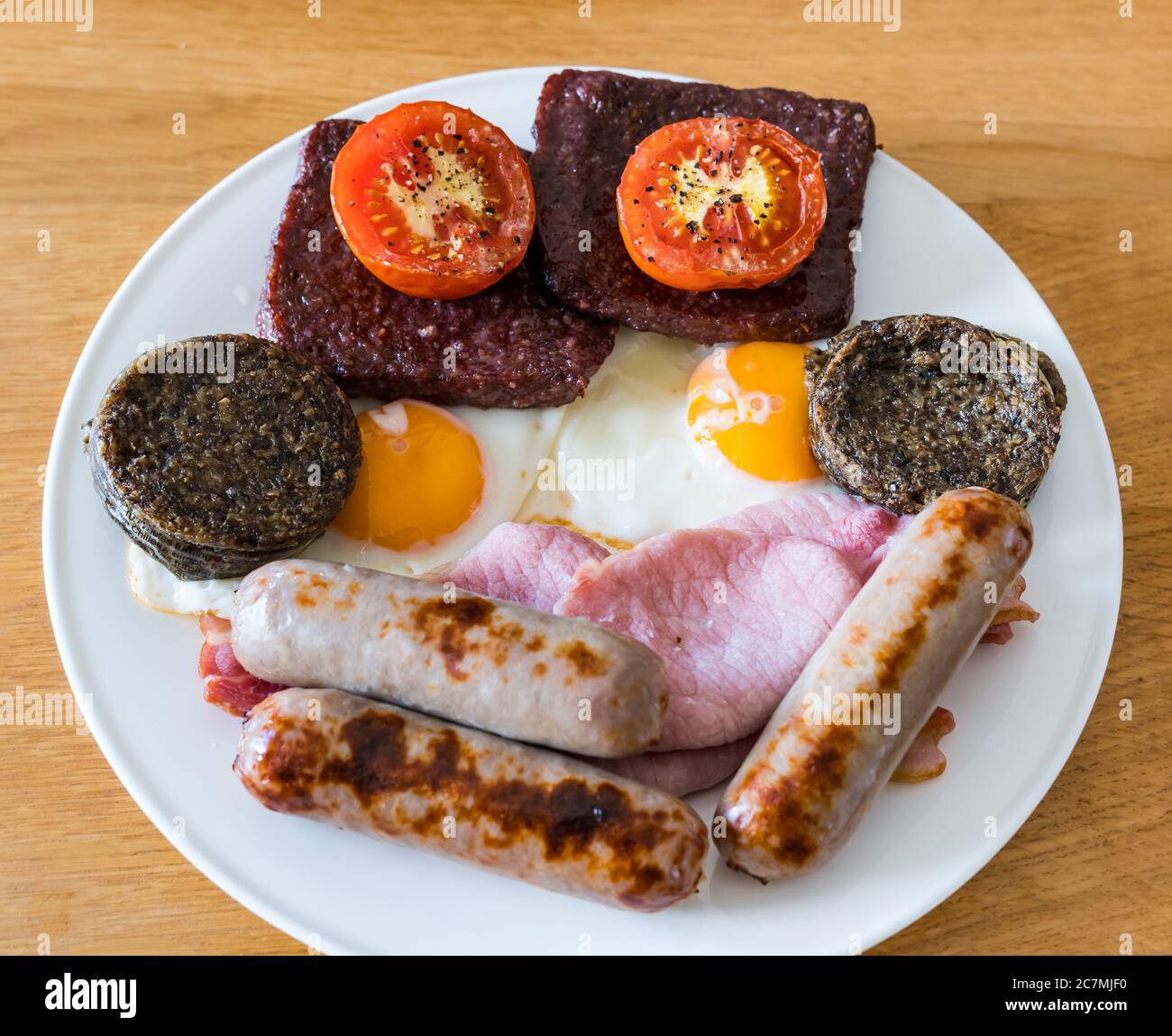 Unhealthy fry up breakfast on white plate with grilled tomatoes, haggis, Lorne sausage meat, pork sausages, fried eggs and bacon Stock Photo
