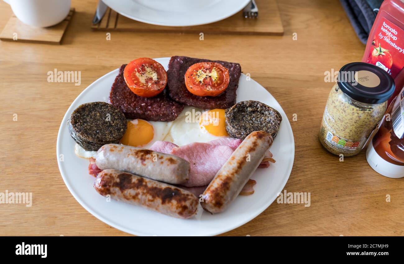 Unhealthy fry up breakfast on plate with grilled tomatoes, haggis, Lorne sausage meat, pork sausages, fried eggs and bacon, mustard and ketchup Stock Photo