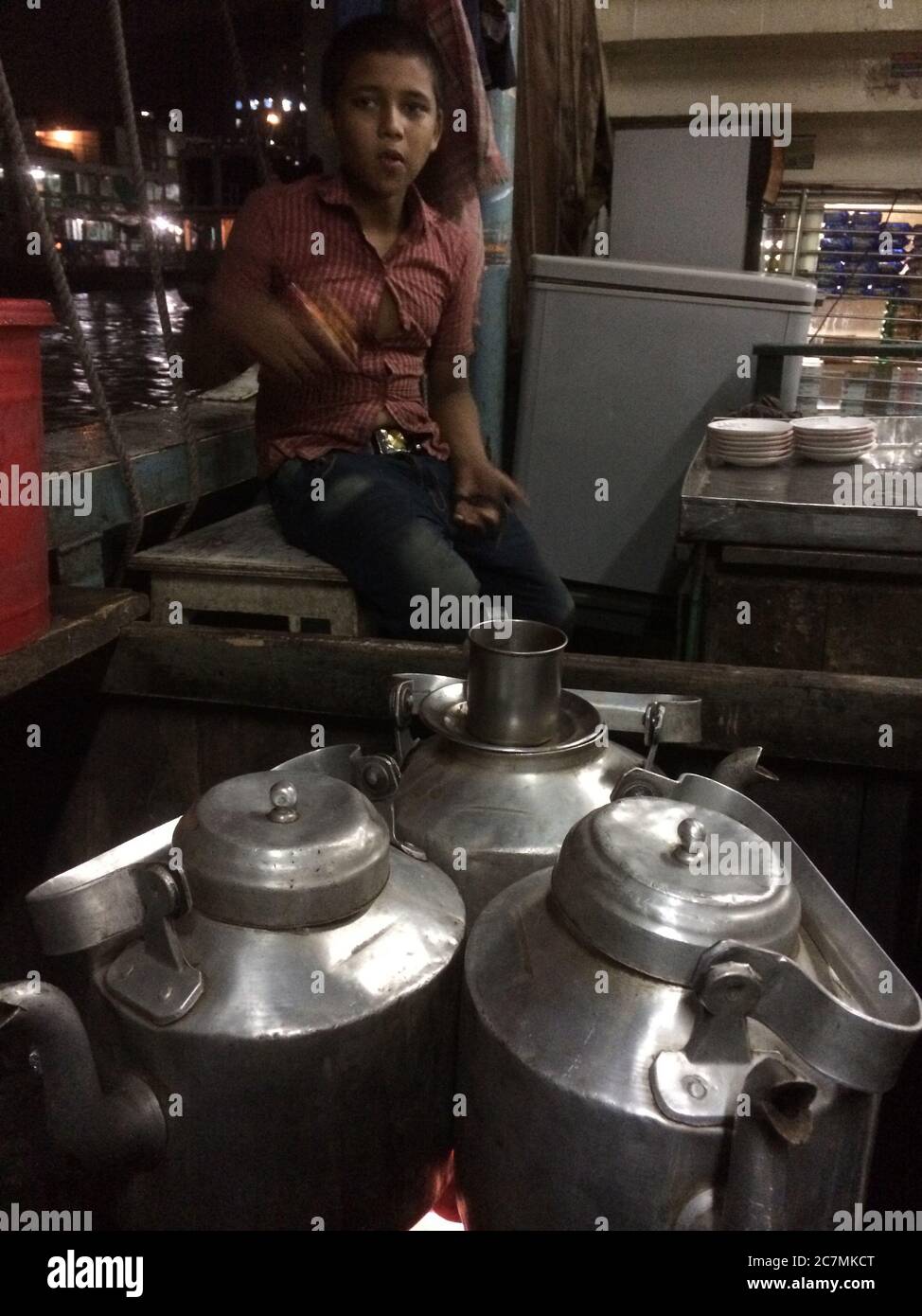 Boy, working a helper in a busy teashop keeps an eye on the boiling tea pots full of hot water in Manikganj, Bangladesh. These open-air shops are constantly serving tea to a string of customers who arrive for refreshment and to catch up on the town gossip. Stock Photo