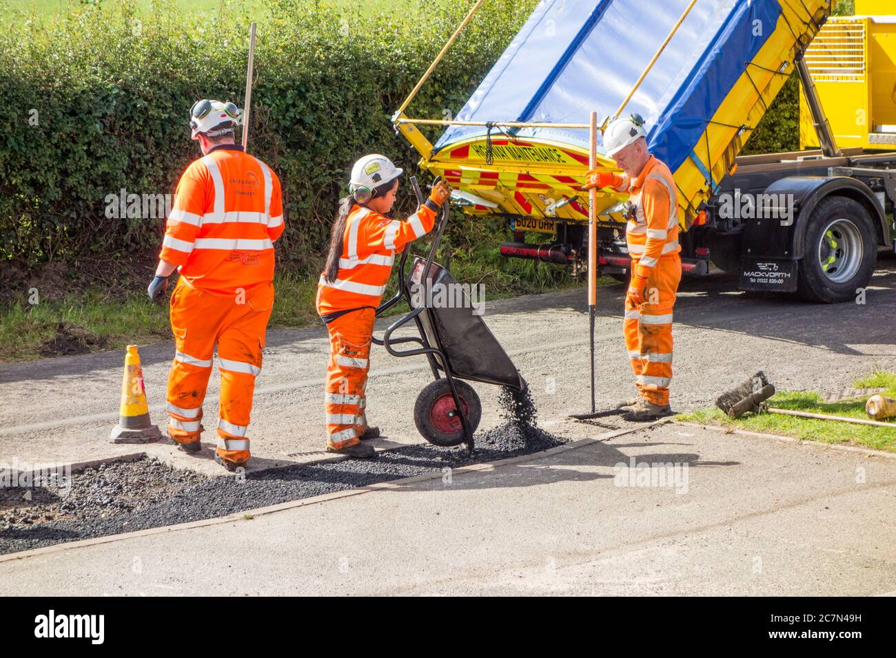 Workmen and woman doing road repairs repairing damaged uneven road surface and pot holes with tarmac in a Cheshire country lane road England UK Stock Photo