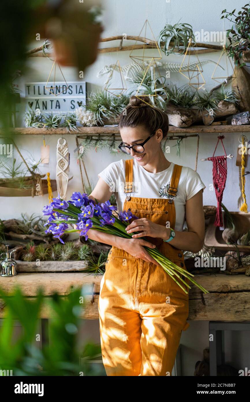 Surprised florist woman in glasses, wear orange overalls surrounded by collection of tillandsia air plants, charmed with pleasant gift, smiling Stock Photo