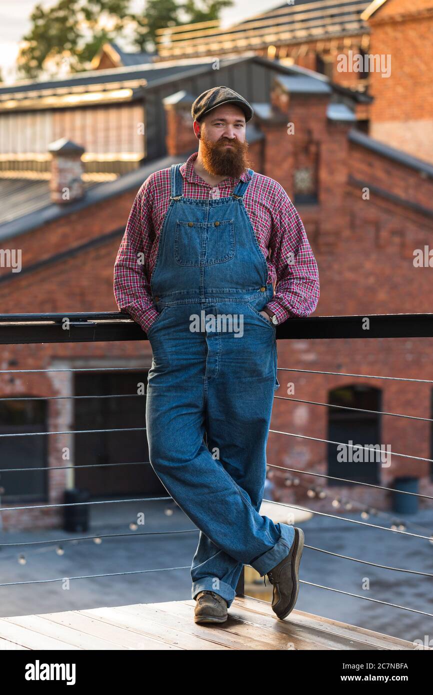 Portrait of brutal bearded motorcycle repairman wearing blue overalls, checked shirt and cap in vintage style of the mid 20th century Stock Photo
