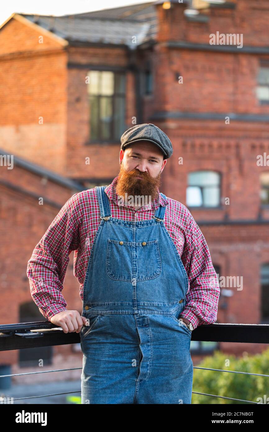 Portrait of brutal bearded man wearing blue overalls, checked shirt and cap in vintage style of the mid 20th century, looking at camera, outdoors. Stock Photo