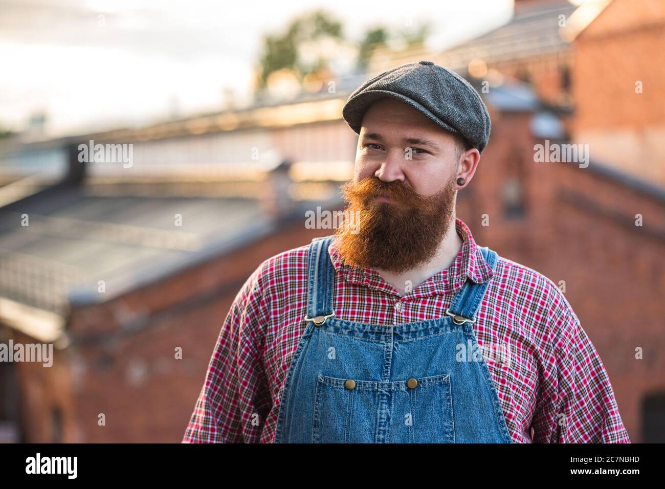 Portrait of brutal bearded stylish craftsman wearing blue overalls, checked shirt and cap in vintage style of the mid 20th century, looking at camera Stock Photo