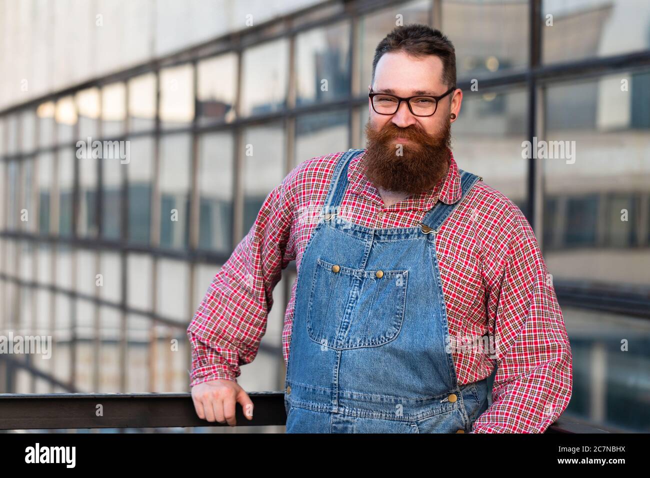 Portrait of brutal bearded stylish craftsman wearing blue overalls, checked shirt in vintage style of the mid 20th century, looking at camera, outdoor Stock Photo