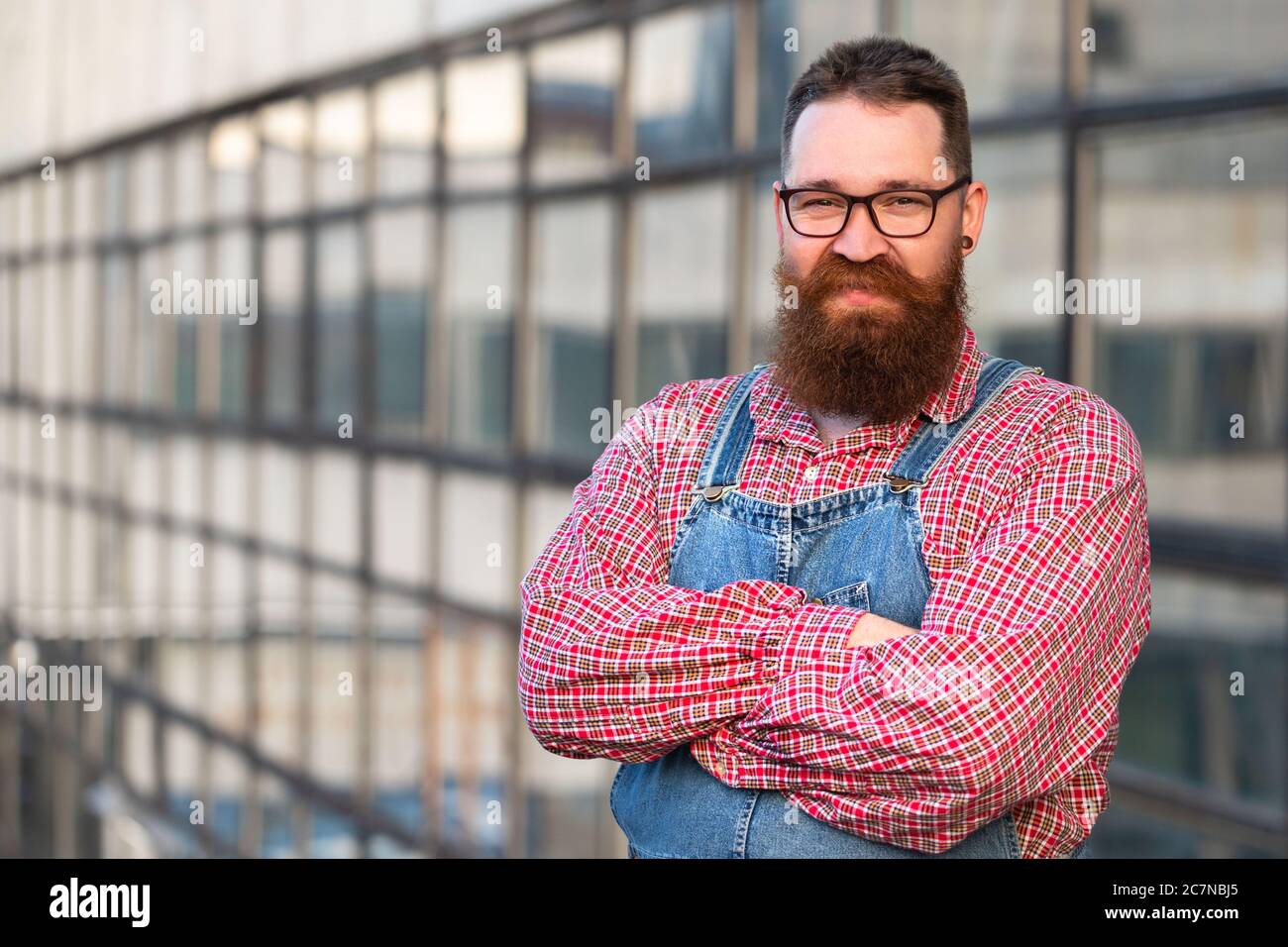 Portrait of brutal bearded stylish craftsman wearing blue overalls, checked shirt in vintage style of the mid 20th century, looking at camera, outdoor Stock Photo