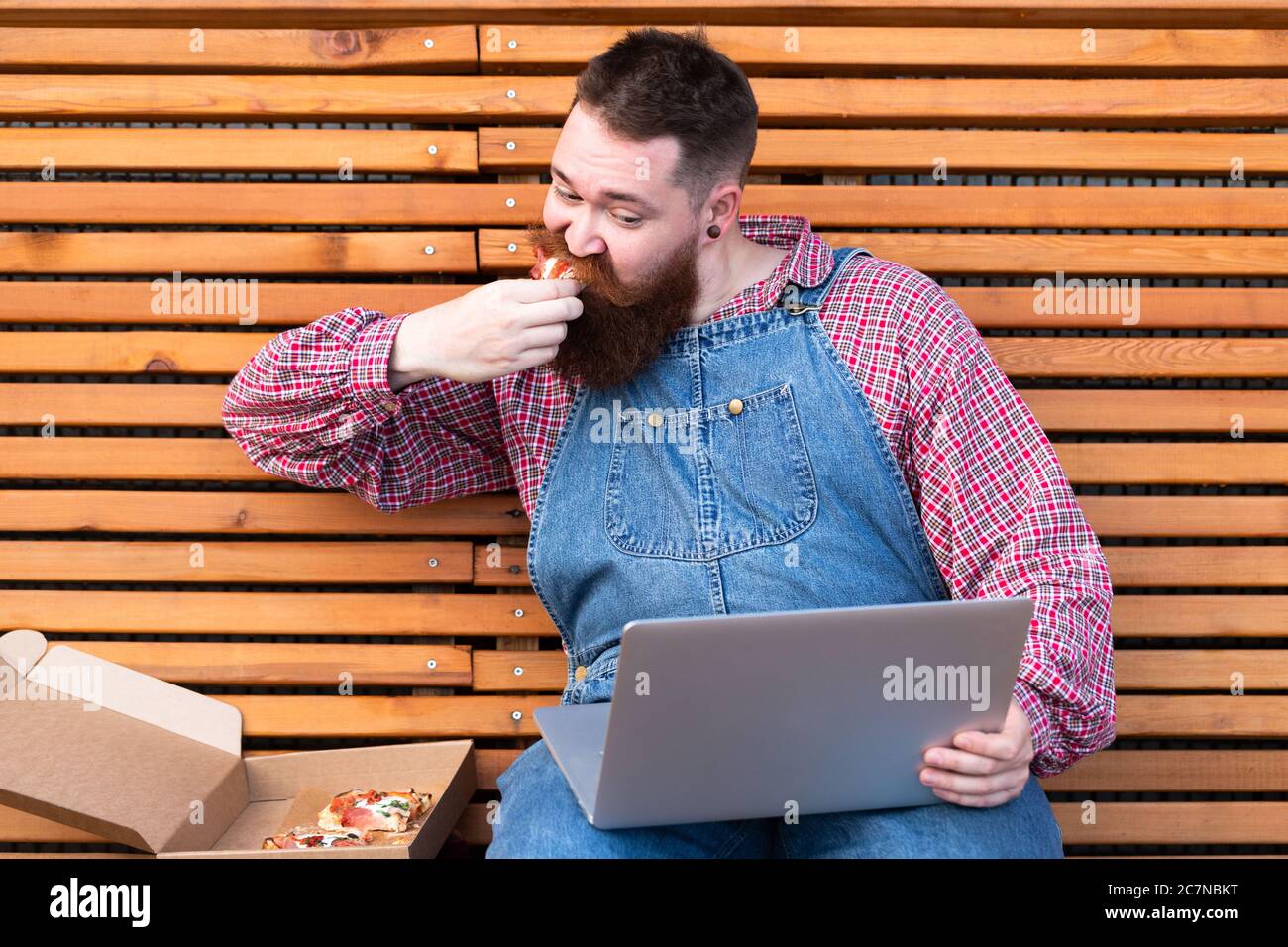 Busy multitask bearded hipster man in blue jeans overalls, checked shirt working on laptop and eating take away pizza, sitting on bench. Fast food Stock Photo