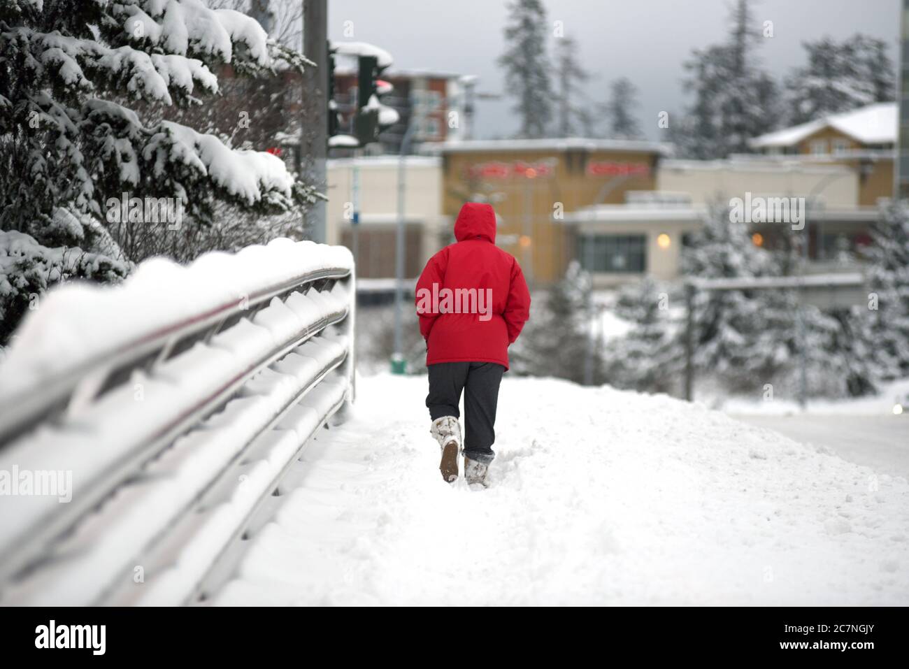 A pedestrian in a red coat makes her way through snow on a sidewalk during a rare west coast snow storm in the greater Victoria suburb of View Royal, Stock Photo