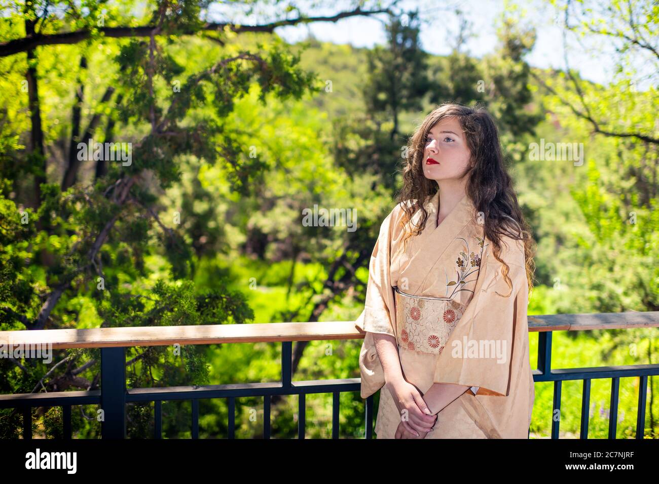 Young caucasian woman dressed up in kimono costume by railing fence in outdoor garden zen home house Stock Photo