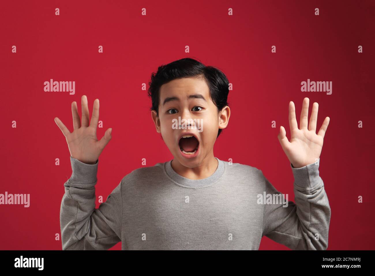 Portrait of funny young Asian boy looking at camera with open mouth and big eyes, shocked surprised expression against red background Stock Photo