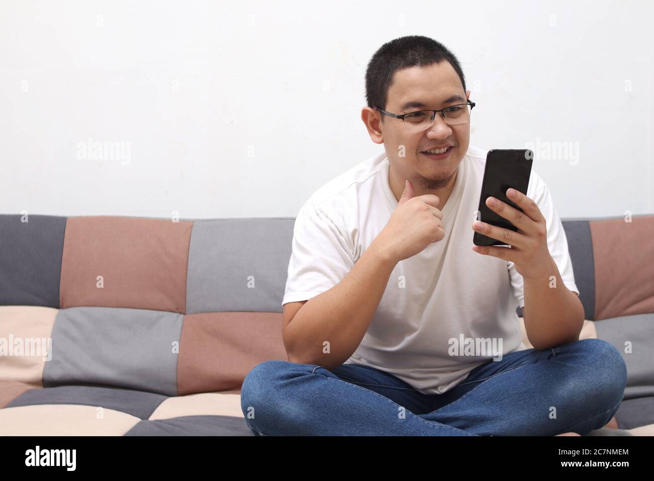 Happy smiling Asian man using smartphone device while sitting on sofa at home. Male reading messages on mobile phone while relaxing at home Stock Photo