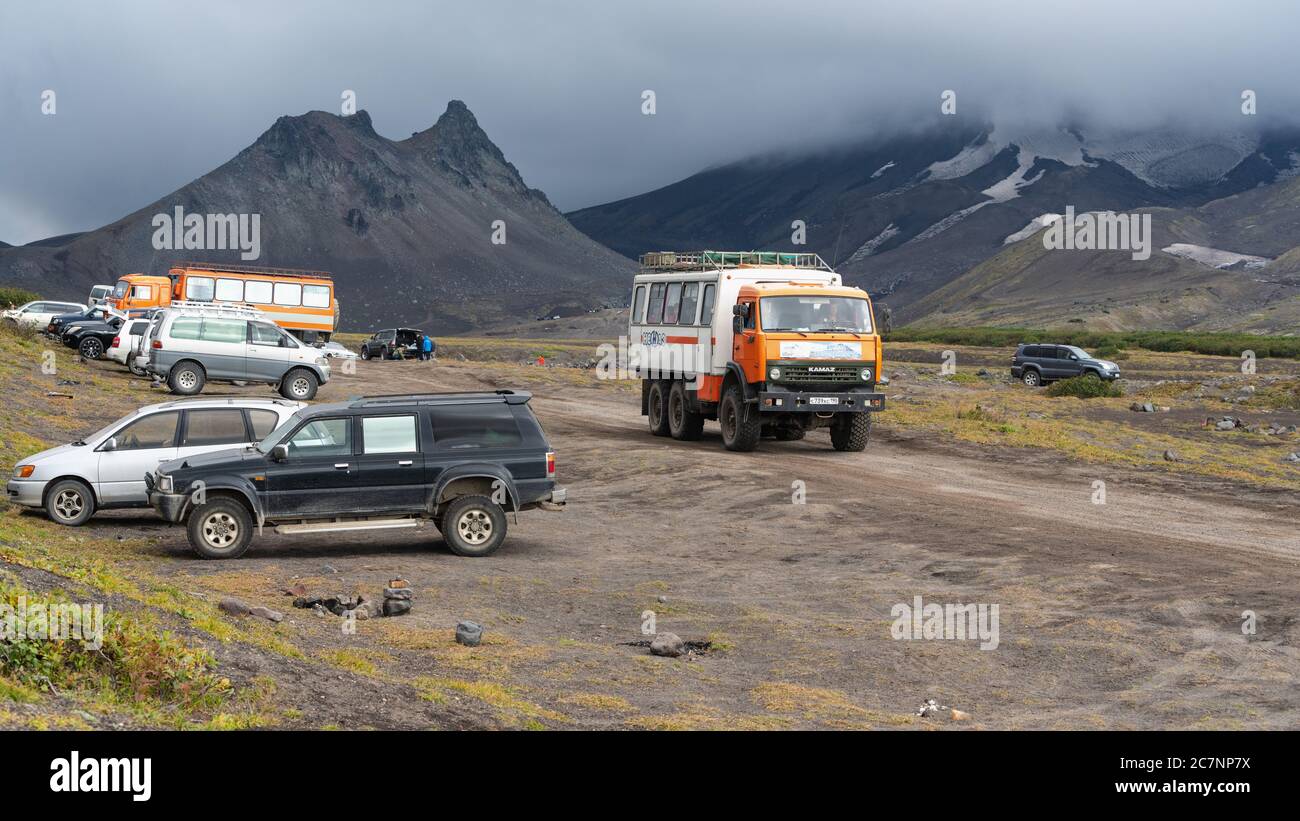 Lots of SUVs and passenger trucks driving and parked along mountain road at foot of volcano in dramatic cloudy weather, volcanic landscape Stock Photo