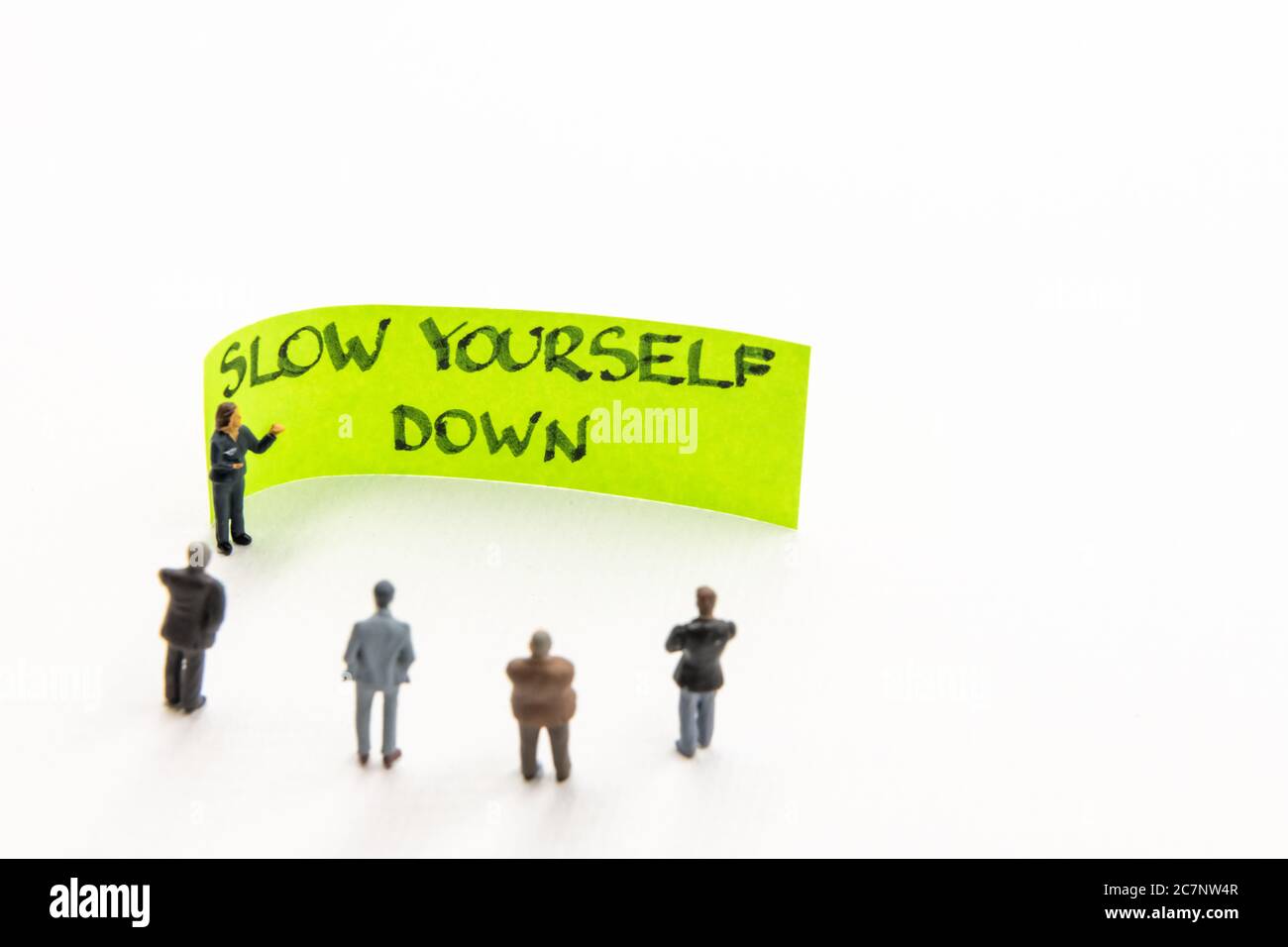 Meeting with miniature figurines posed as business people standing around post-it note with Slow Yourself Down handwritten message on white background Stock Photo