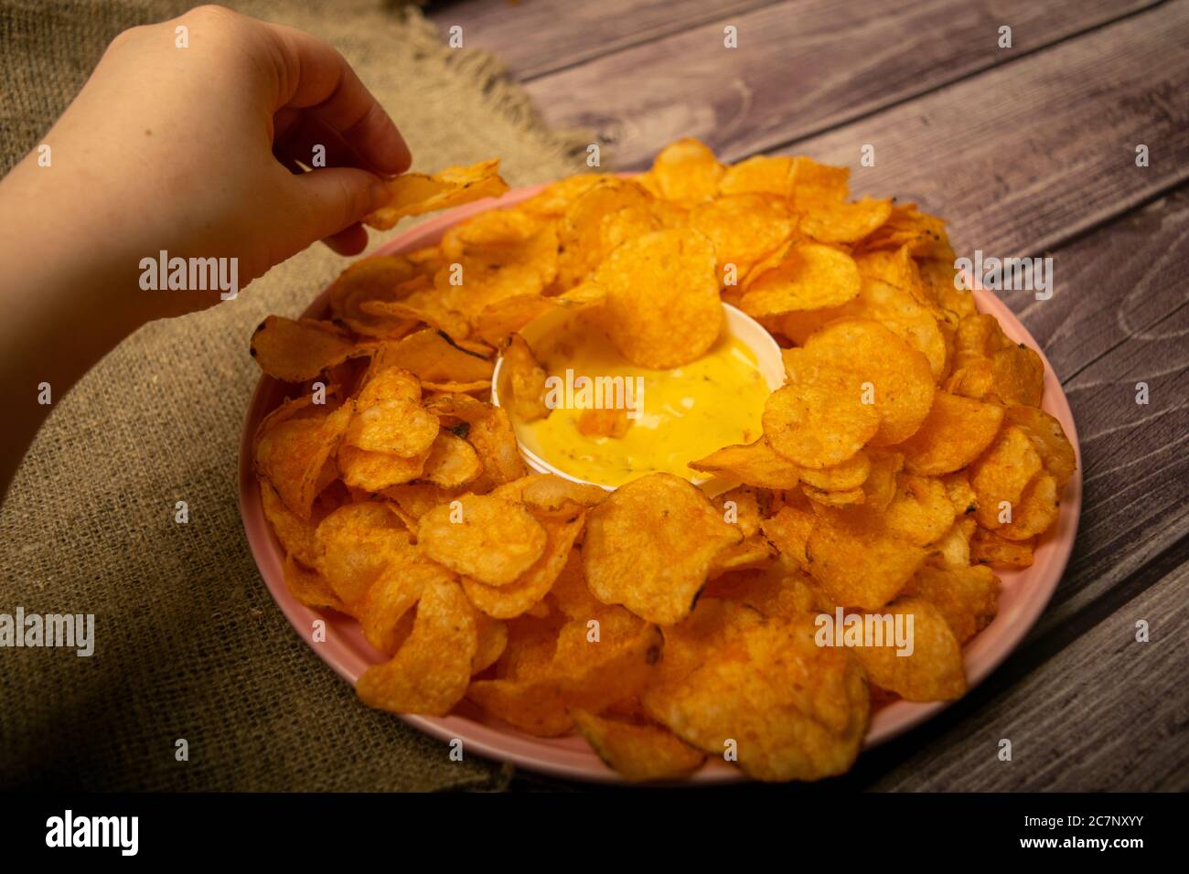 The girl takes a chip from a round dish with potato chips and a saucepan with cheese sauce in the center of the plate. Close up Stock Photo