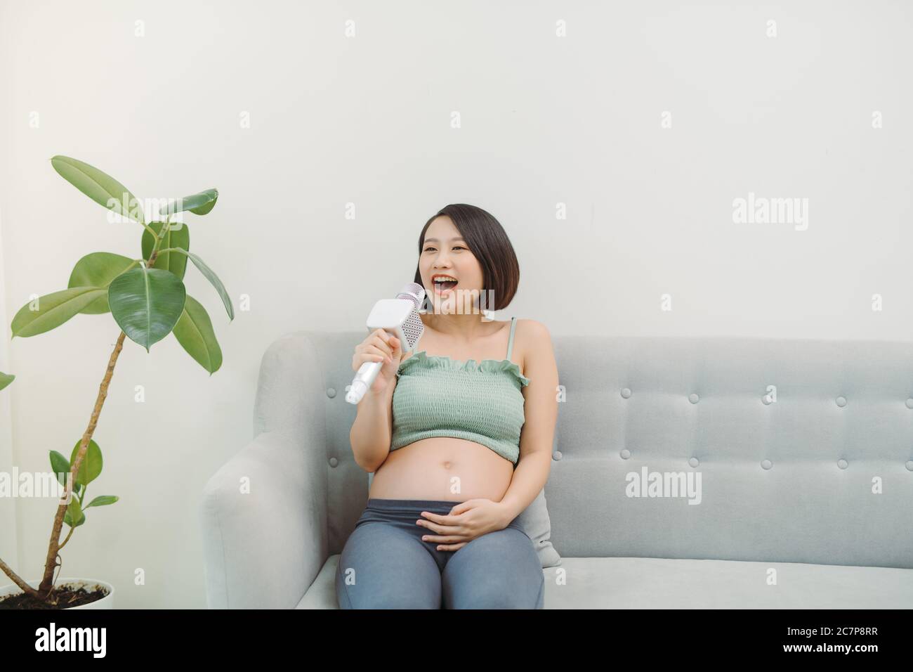 Beautiful young pregnant woman having fun: asian holding microphone and singing a song Stock Photo