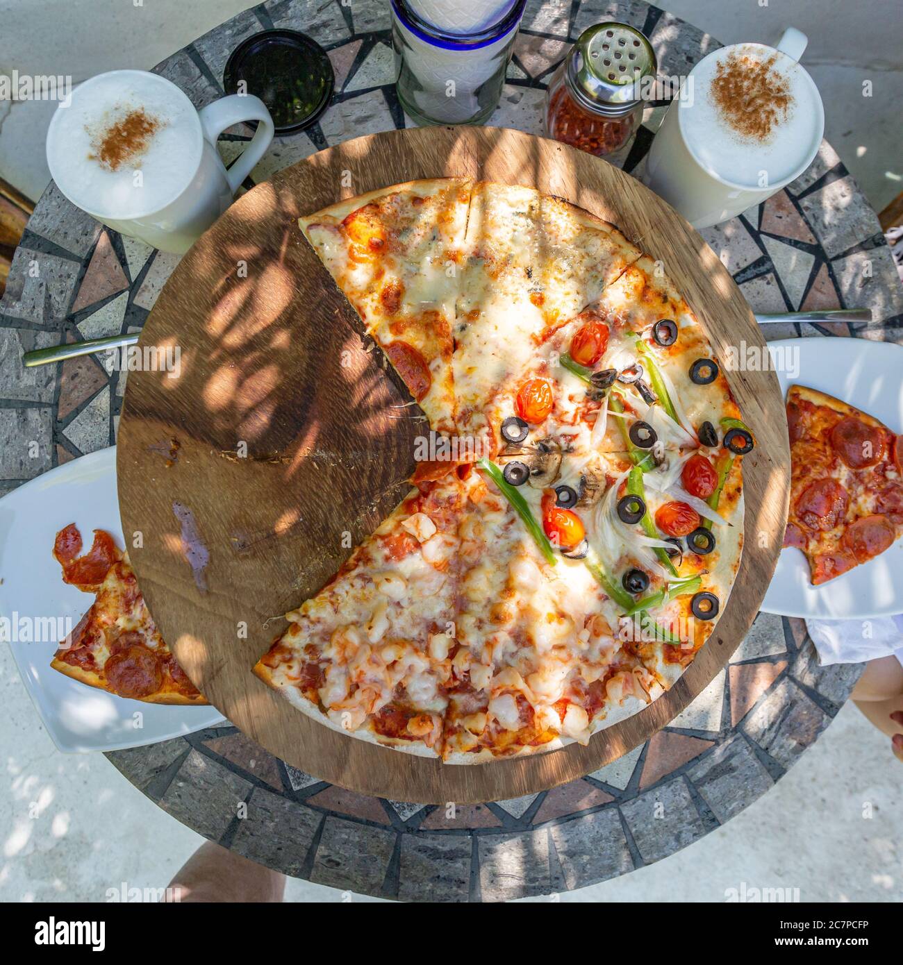 Top view of a four season pizza on a stone table with a wooden base next to two cups of cappuccino coffee, plates with half-eaten pizza slices Stock Photo