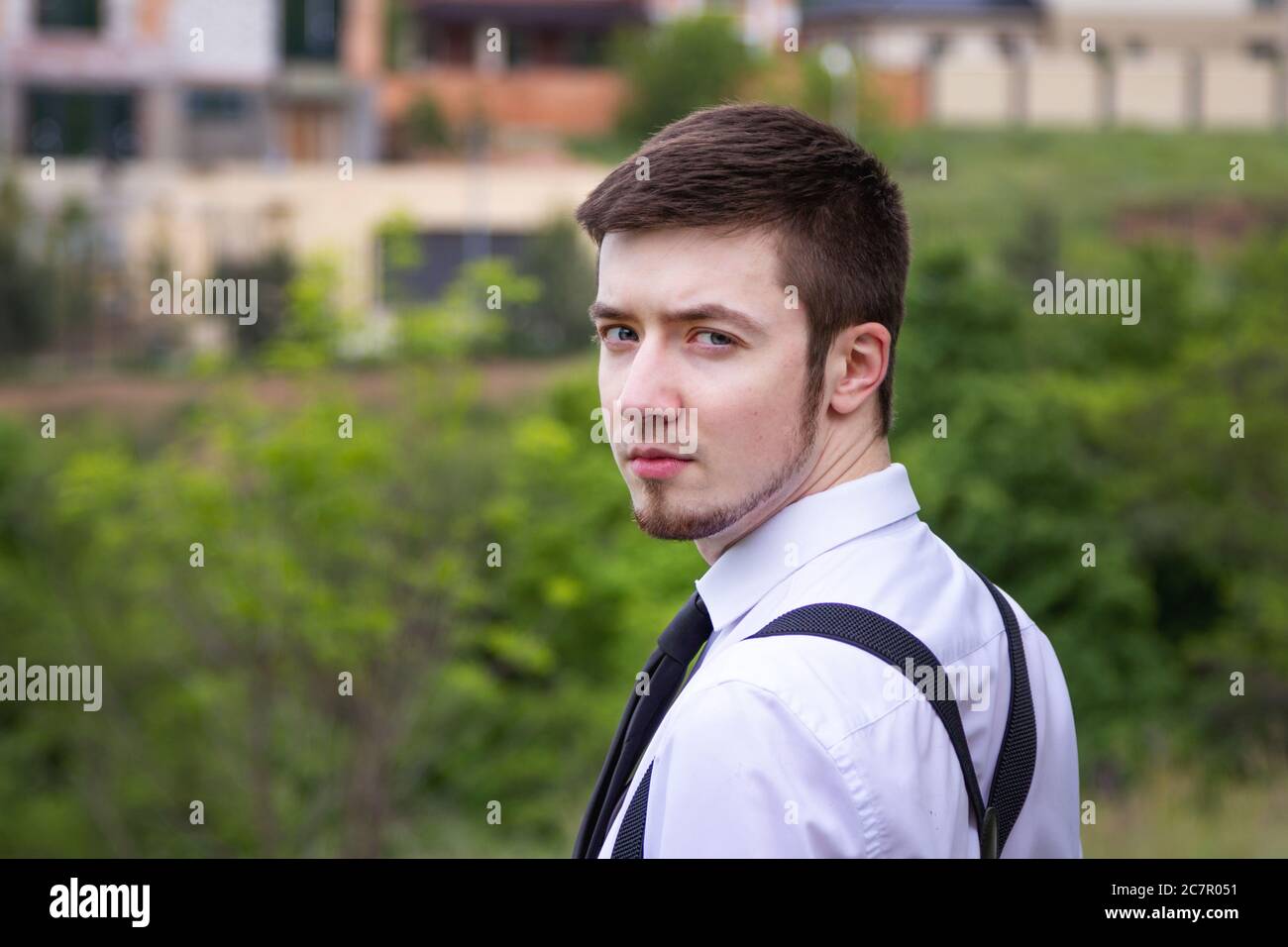 Portrait of stylish brunet young man, wearing white shirt, black tie, suspenders and glasses, walking on the meadow near river. Stock Photo