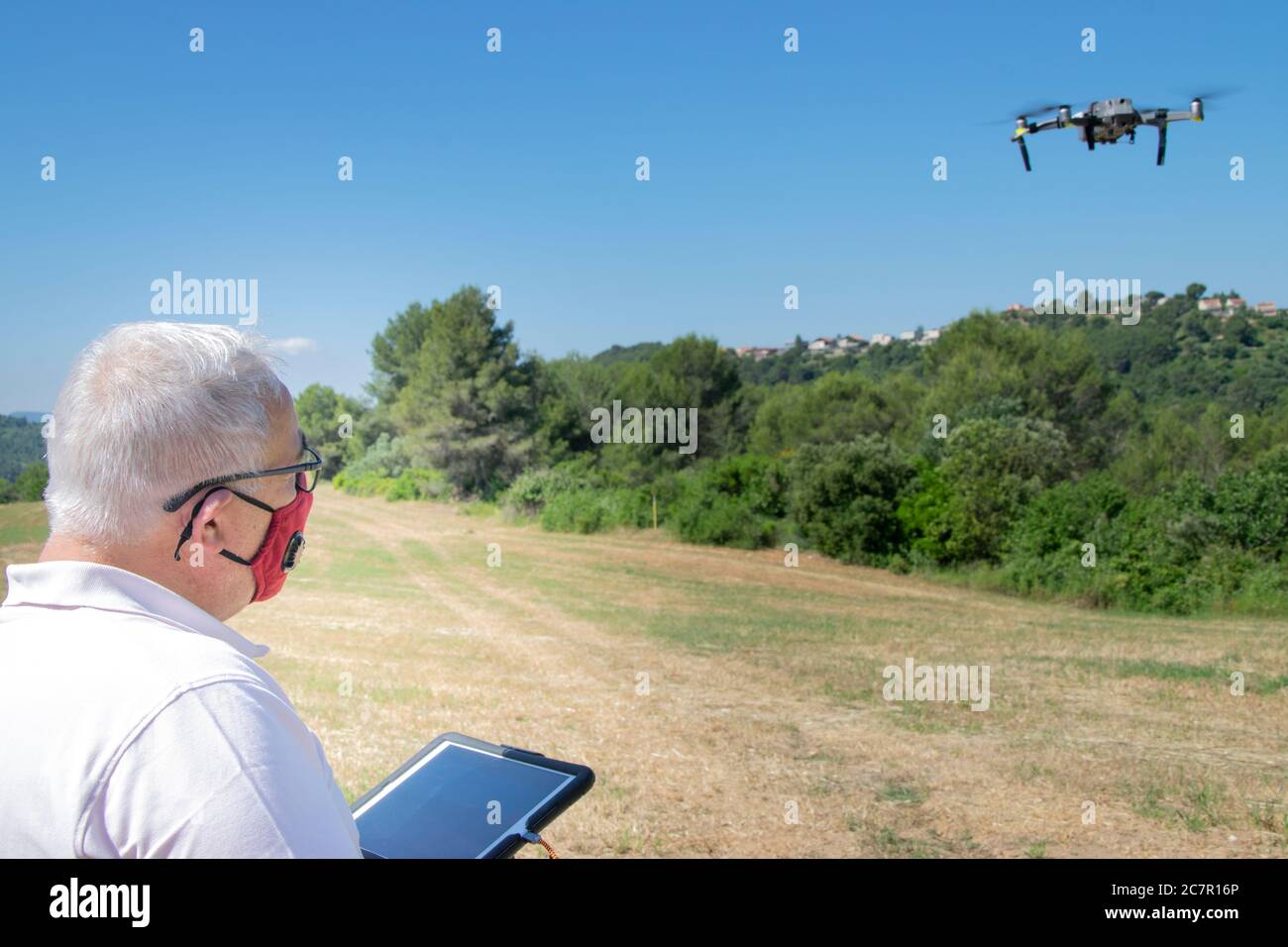 Man with grey hair flying a drone on a field with a tablet and a safety face mask. New normality concept. Stock Photo