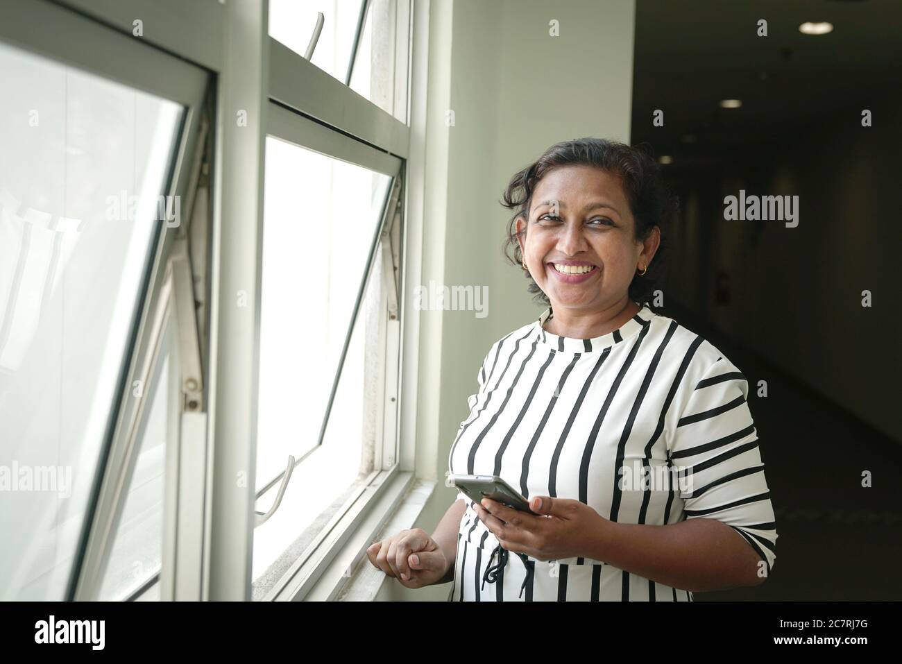 Indian businesswoman standing next to windows, holding cellphone and smiling at the camera Stock Photo