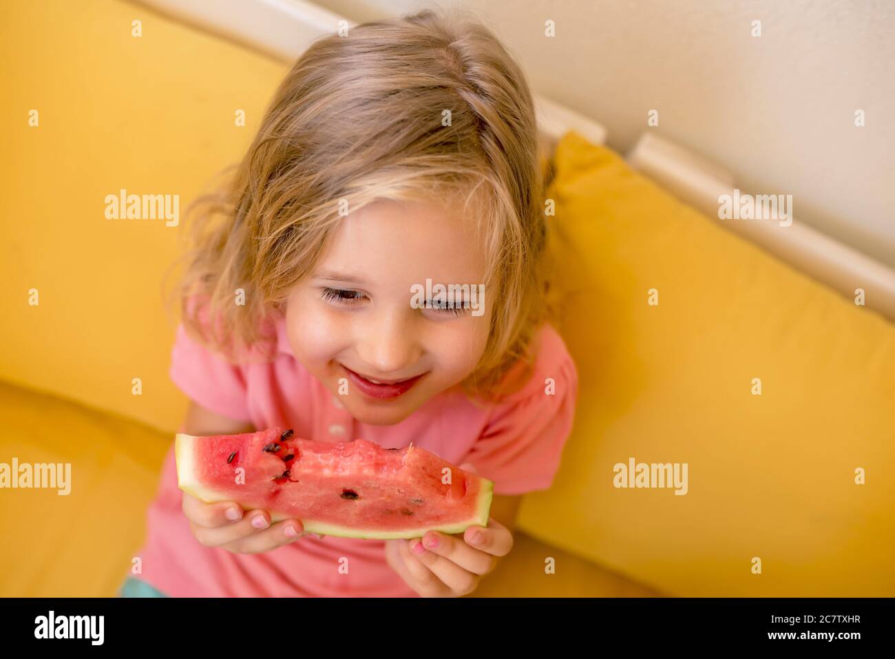 High angle closeup shot of a female child smiling and eating a watermelon on a yellow couch Stock Photo