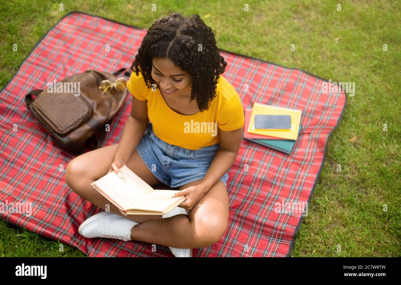 Pretty black girl reading book on picnic blanket at park, above view. Free space Stock Photo