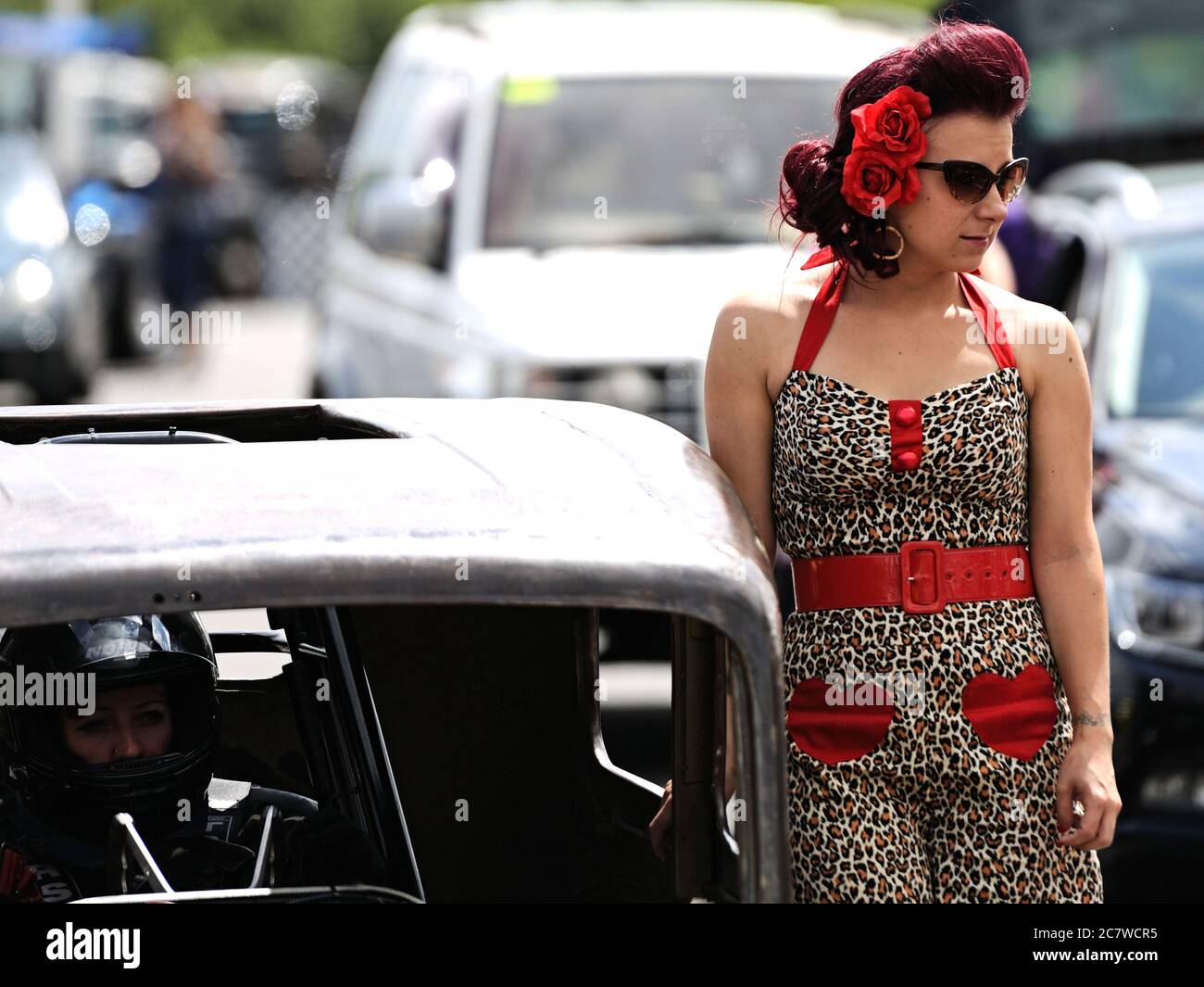 A girl in a Fifties period dress, wearing sunglasses and red flowers in her hair stands next to a dragster at Santa Pod Raceway Stock Photo
