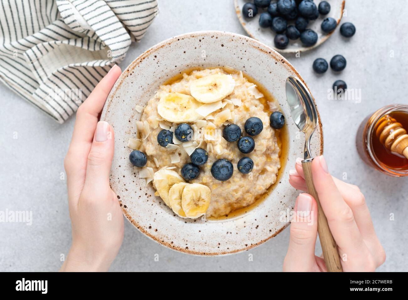 Oatmeal porridge with banana, blueberries and honey. Female hands holding bowl of oatmeal porridge over grey concrete background, table top view Stock Photo