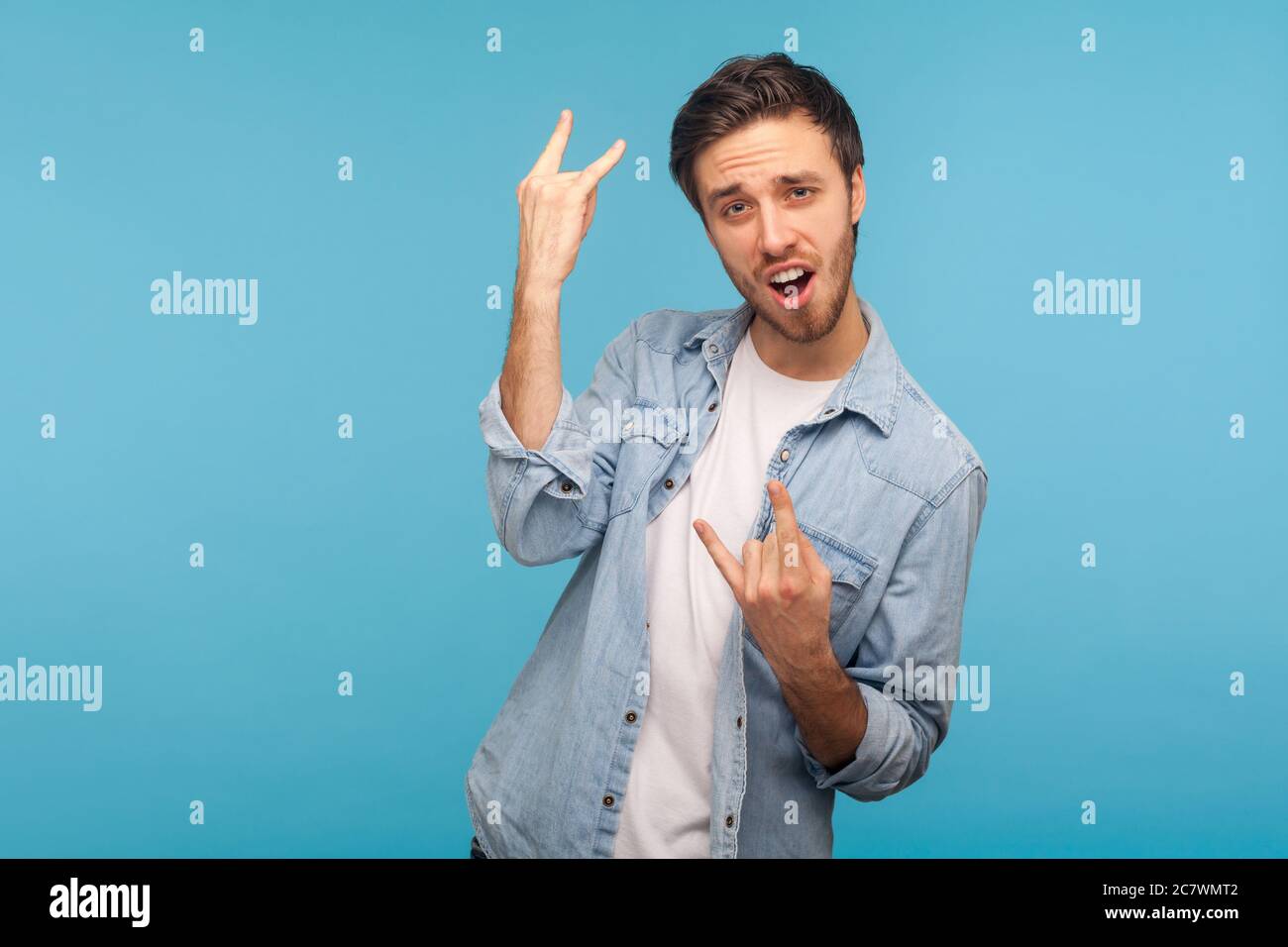 Yeah, that's crazy! Portrait of enthusiastic man in worker denim shirt showing rock and roll hand gesture and screaming in delight, making devil horns Stock Photo