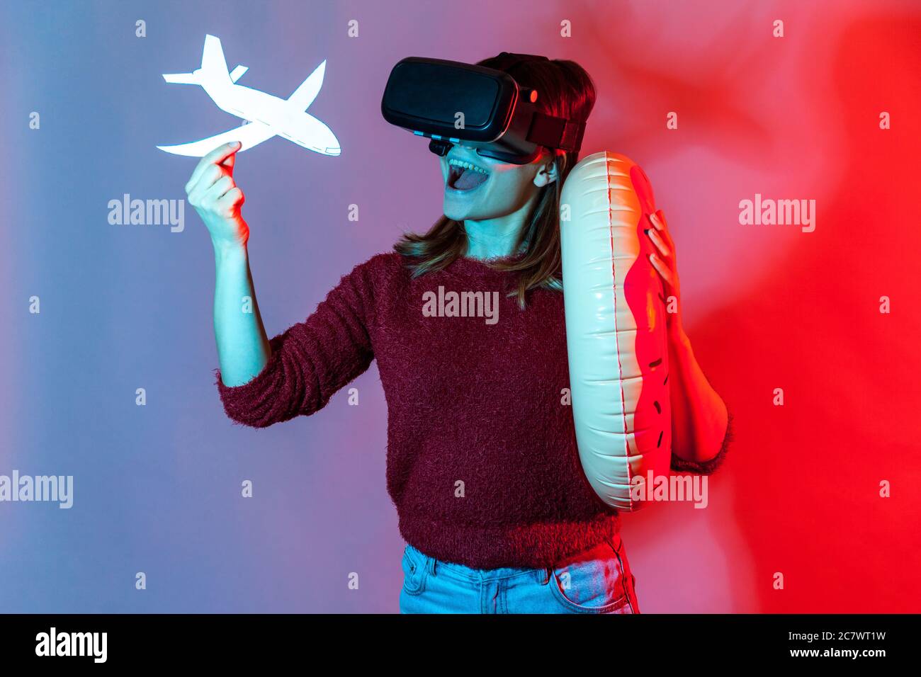Joyful tourist girl in VR glasses holding rubber ring and airplane mock up, watching virtual travel tours, vacation in tropical country, having journe Stock Photo
