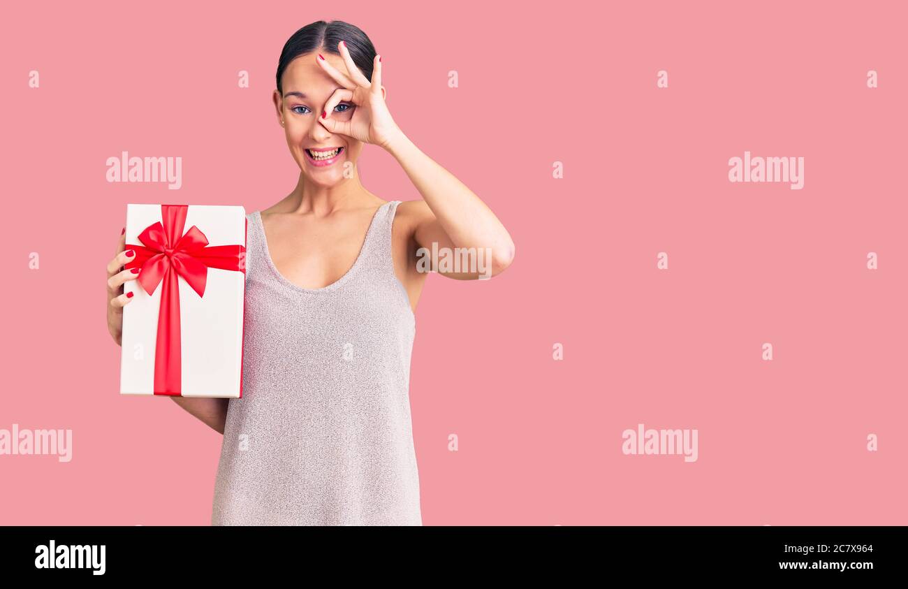 Beautiful brunette young woman holding gift smiling happy doing ok sign with hand on eye looking through fingers Stock Photo