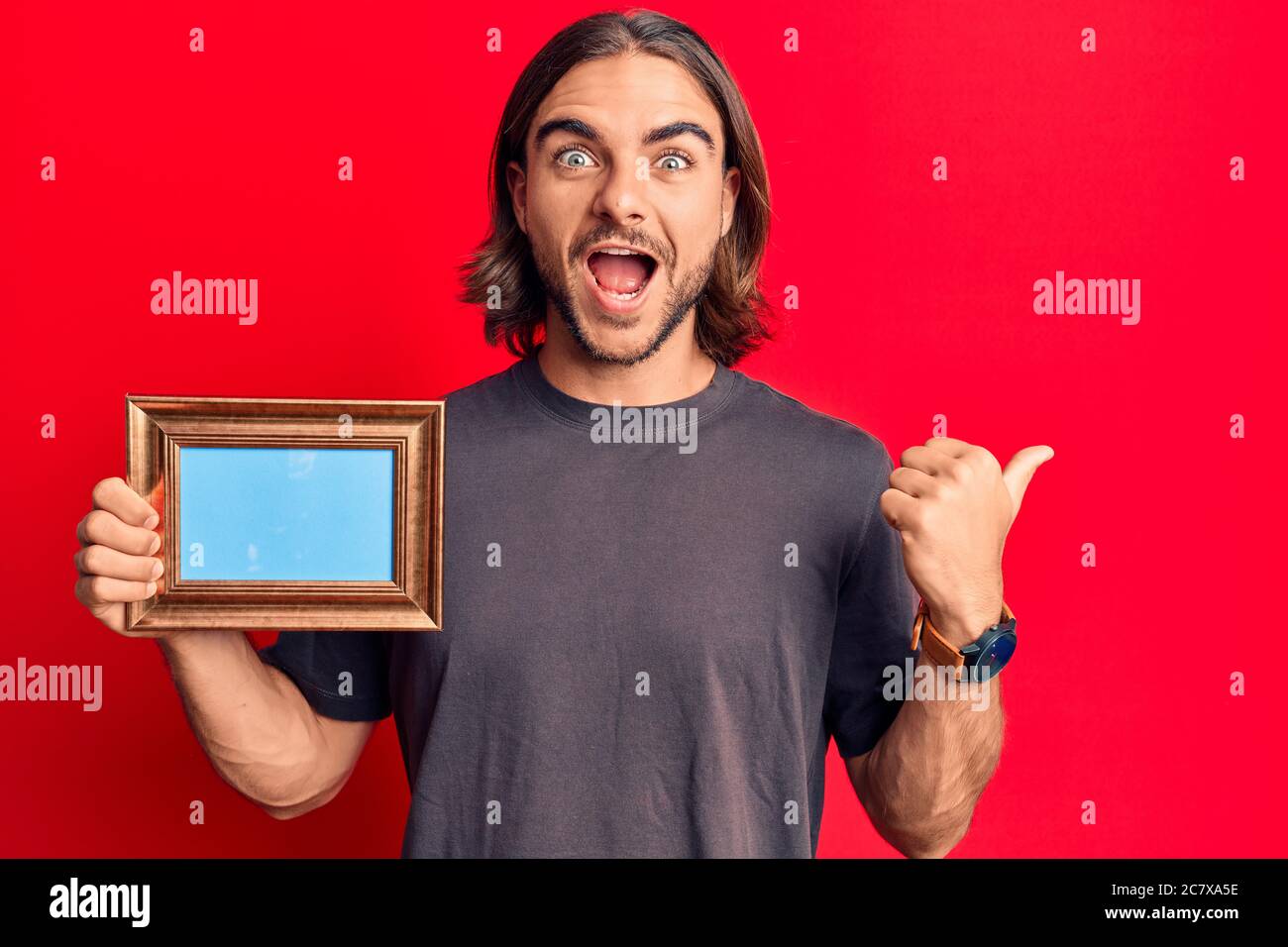 Young handsome man holding empty frame pointing thumb up to the side ...