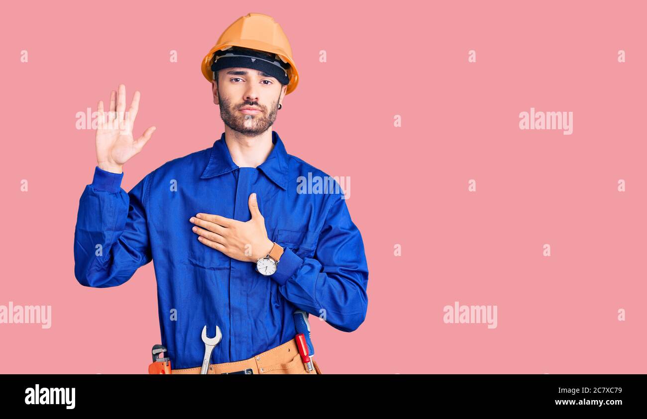 Young hispanic man wearing worker uniform swearing with hand on chest ...