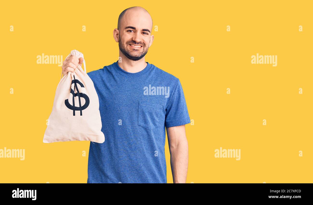 Young handsome man holding money bag with dollar symbol looking positive and happy standing and smiling with a confident smile showing teeth Stock Photo