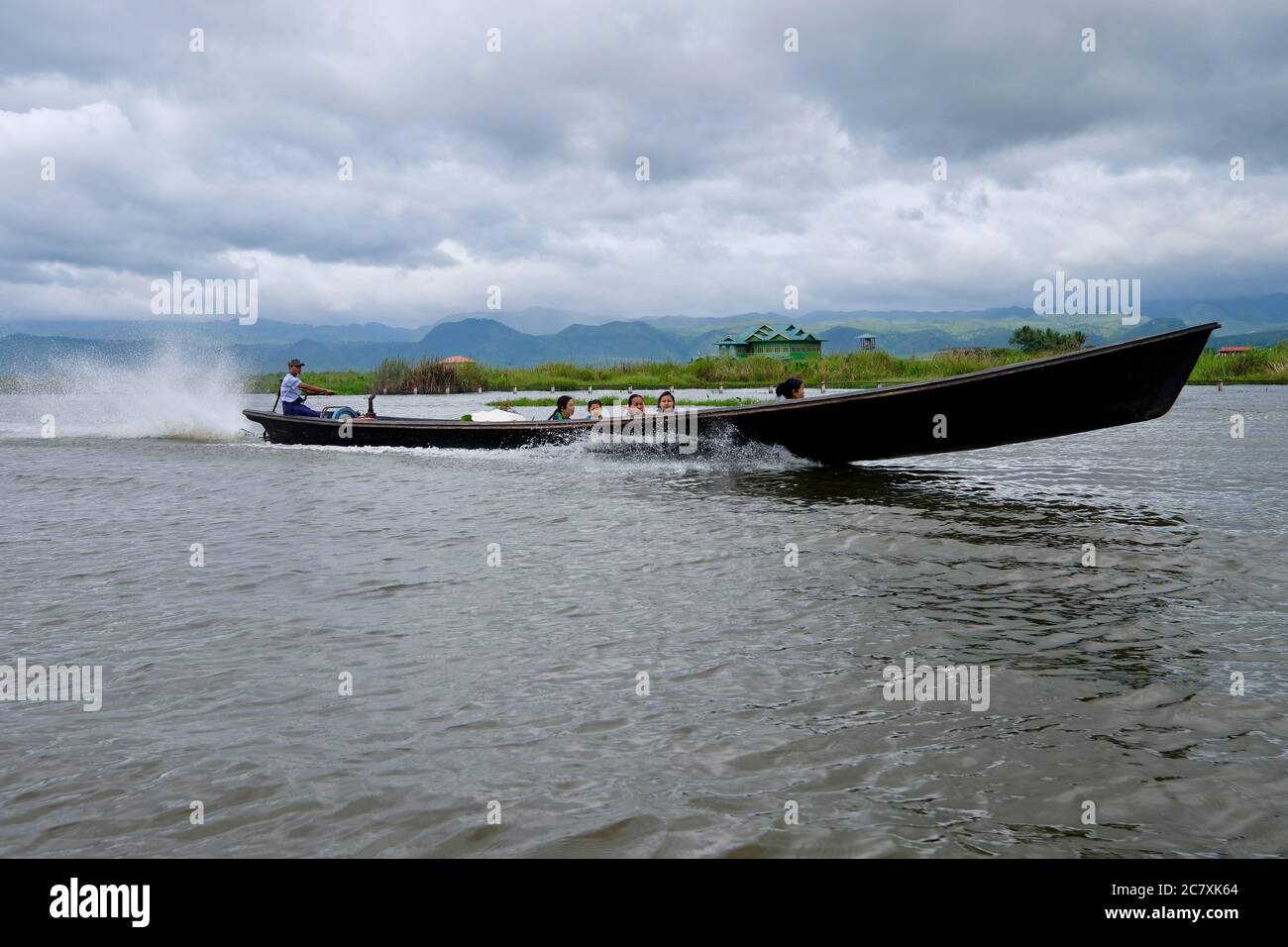 Burmese family driving a boat on Inle lake. Green plants and house above water. White cloudy sky Stock Photo