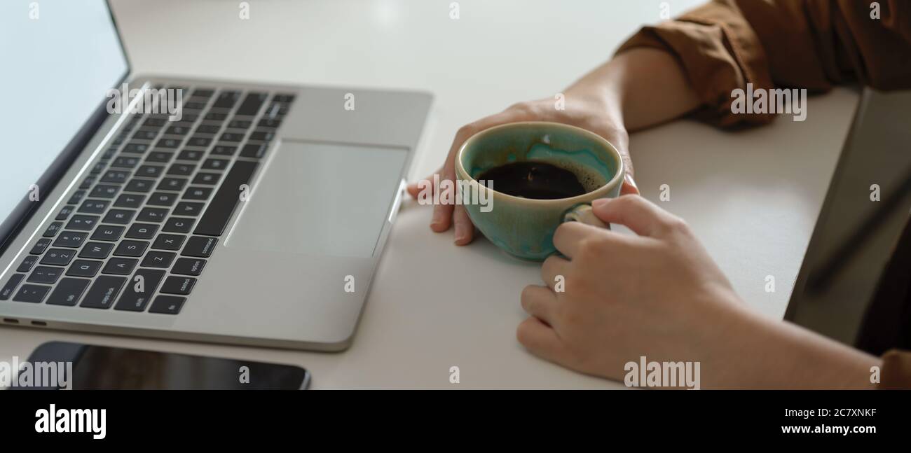 Side view of female holding coffee cup while looking on mock up laptop on white table with smartphone Stock Photo